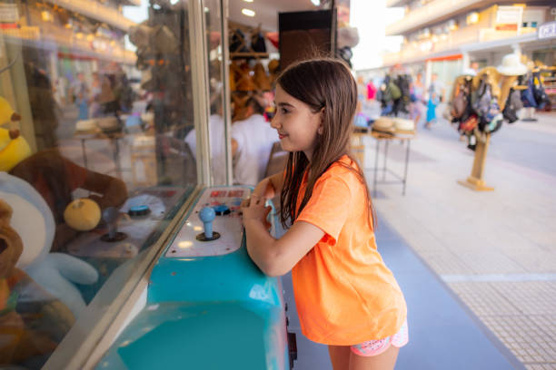 Adorable girl in orange reaches for toys in a claw machine at the arcade.